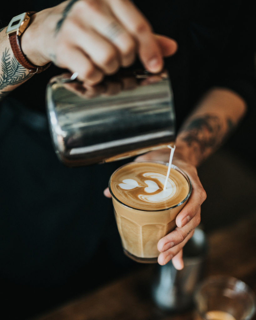 Een getatoeëerde hand giet melk in een glas en creëert vakkundig een hartvormig ontwerp op het oppervlak van een latte voor de promotiecampagne van een online marketeer.