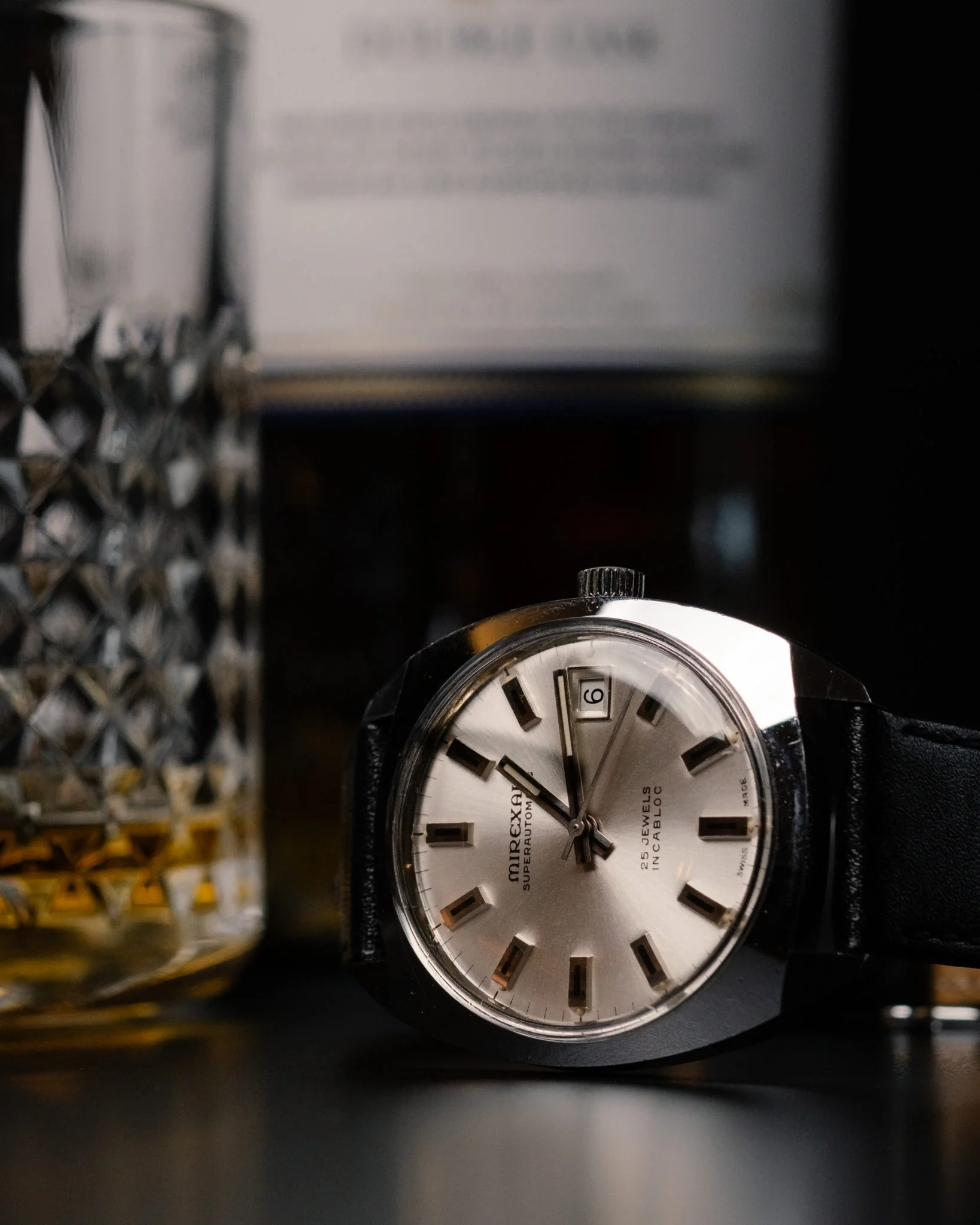 Een close-up van een Mirexal superautomatisch polshorloge met een zilveren wijzerplaat, zwarte band en een wazige kristallen karaf op de achtergrond, wat een elegante setting suggereert.