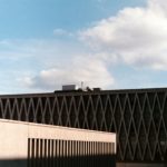 De afbeelding toont het bovenste gedeelte van een modern gebouw met geometrische patronen op de gevel, onder een heldere hemel met minimale bewolking.