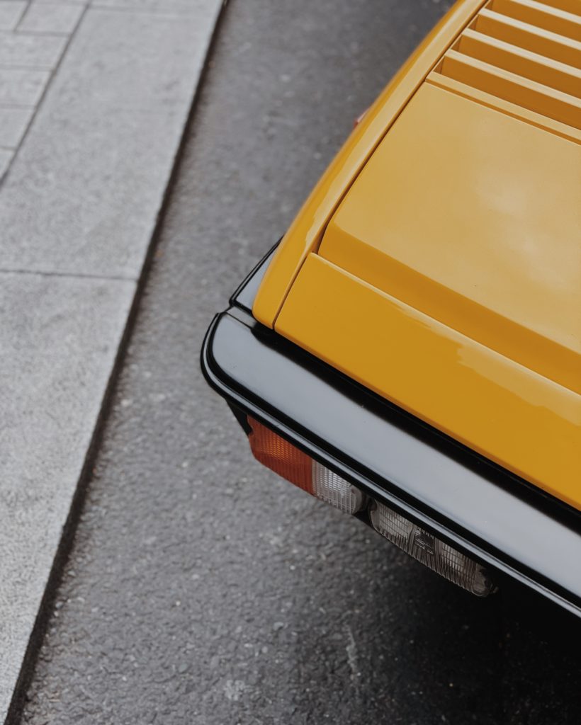 Een close-up van de achterkant van een gele auto, waarbij de nadruk ligt op het achterlicht en de kofferbak naast een grijze stoep.
